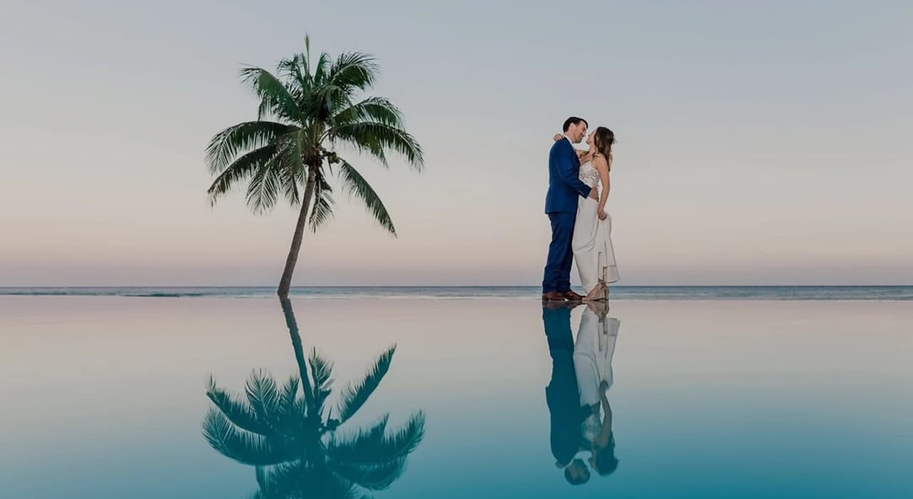 Pareja de novios frente al mar, reflejados en la piscina infinita al atardecer